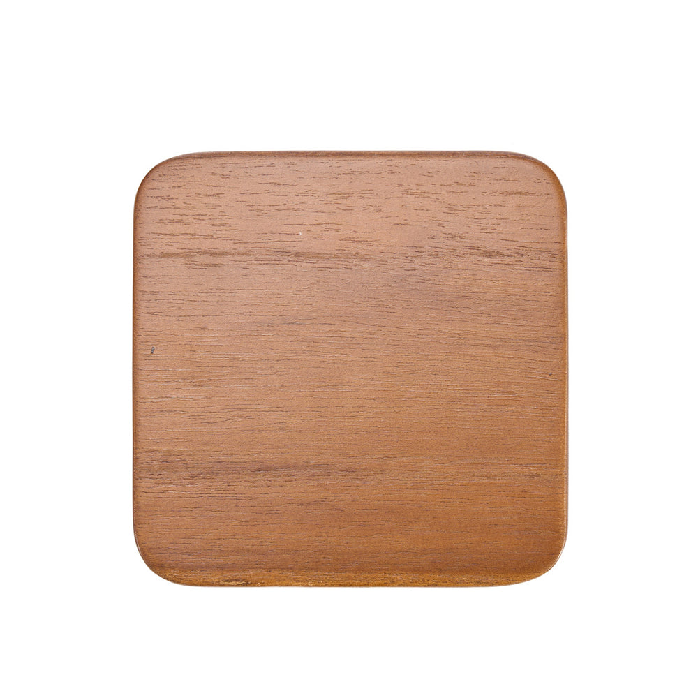 A medium brown wood coaster rests quietly on the table, its smooth teak surface catching soft light and revealing subtle grain and warm, grounded tone.